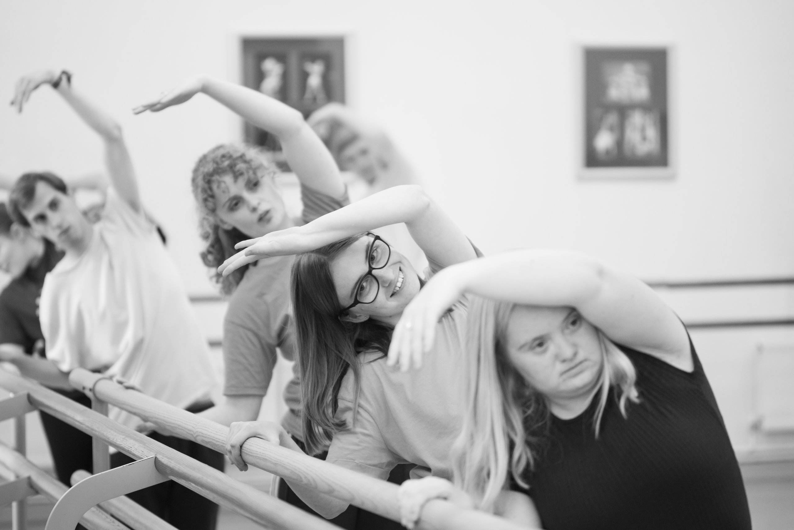 3 people leaning towards a ballet barre with one arm curving over their heads and the other holding the ballet barre.